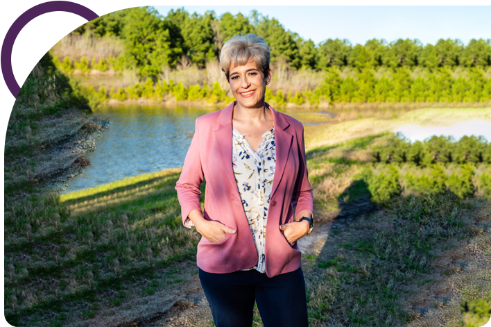 A woman in pink jacket standing on grass near water.